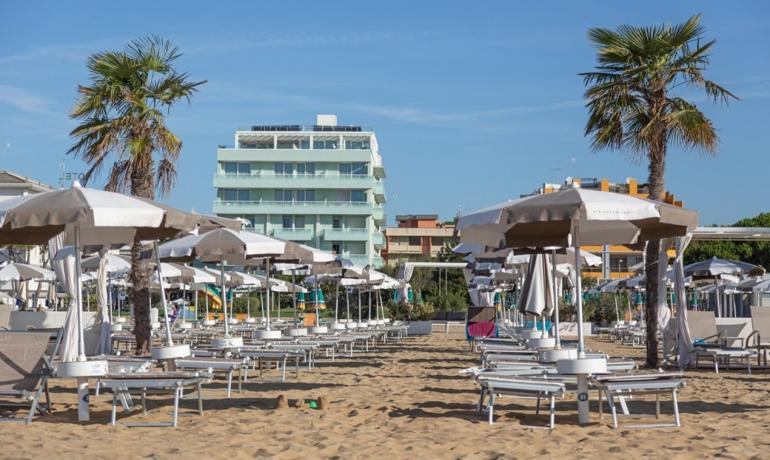 Beach with umbrellas and sunbeds, palm trees and buildings in the background.