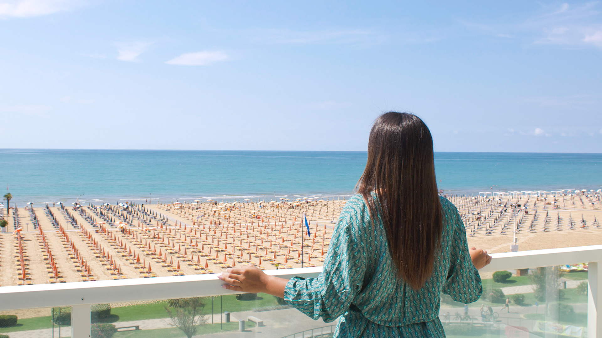 Woman looks at the sea from a balcony with beach and umbrellas.